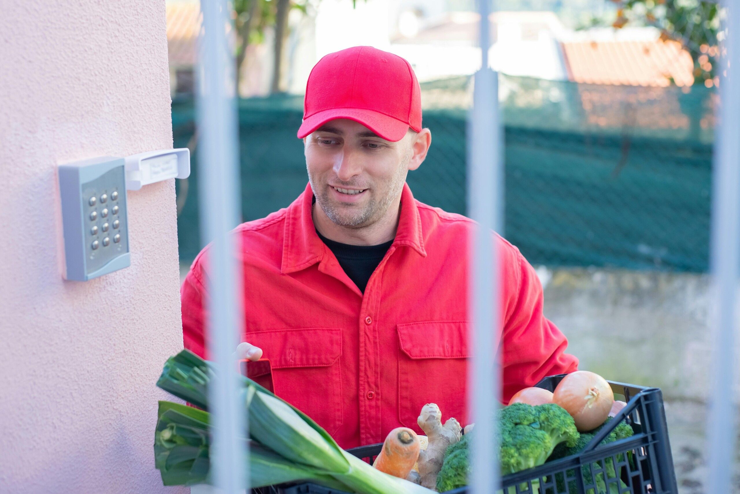 Una persona con una camisa roja y una gorra roja está sosteniendo una caja de verduras frente a un teclado de seguridad en una pared.